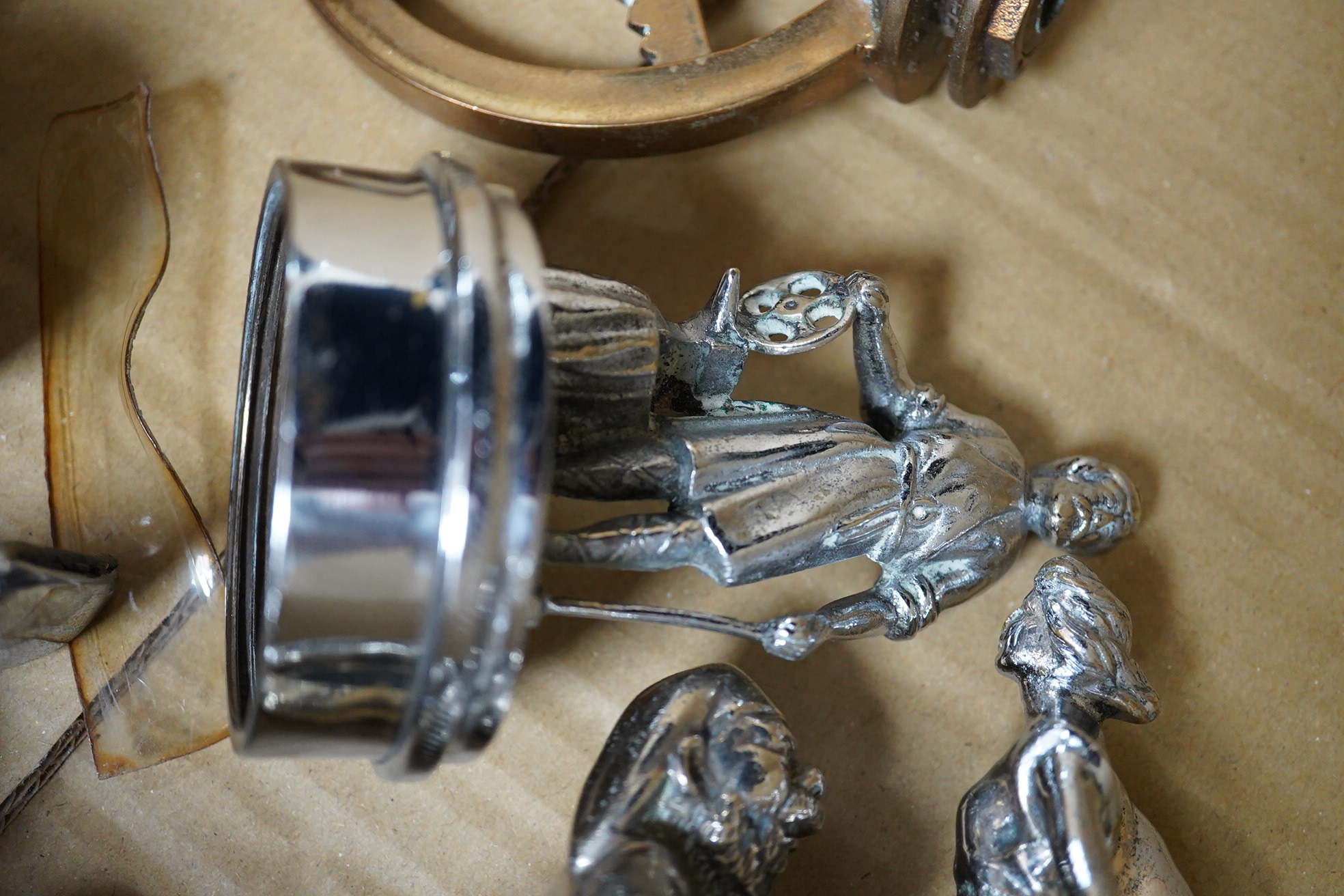 Seven chrome and brass car mascots, including a Mercedes-Benz logo, a figure of a blacksmith, a caricature figure on a beer barrel, a golf ball in mid flight, etc. together with a chromed housing unit for an electric lam